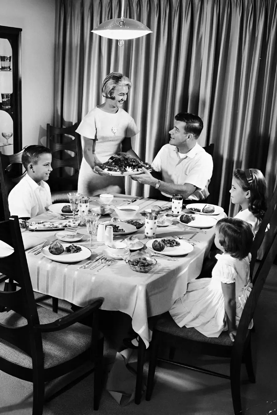 A smiling woman serves a platter of food to a seated man at a dining table with two girls and a boy. The family is dressed neatly, sharing a meal in a warmly lit, mid-century dining room.