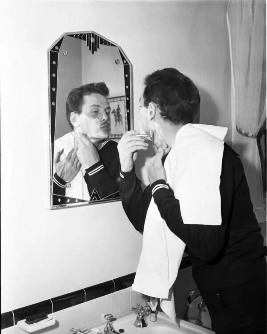 A man stands at a bathroom sink, shaving his face while looking into a mirror. He has a towel draped over his shoulder and uses one hand to stretch his skin and the other to hold a razor.