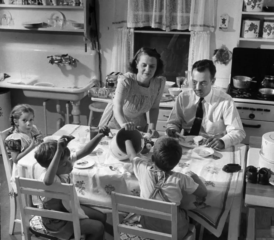 A family of four, two adults and two children, sits around a kitchen table enjoying a meal together. The mother serves food while the father and kids sit, with a stove and sink visible in the background.
