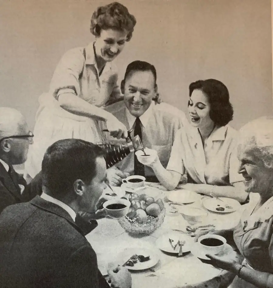 Five adults sitting around a table, smiling as a woman pours coffee. The table has cups, saucers, and a bowl of fruit. The scene appears cheerful and social, resembling a 1950s gathering.