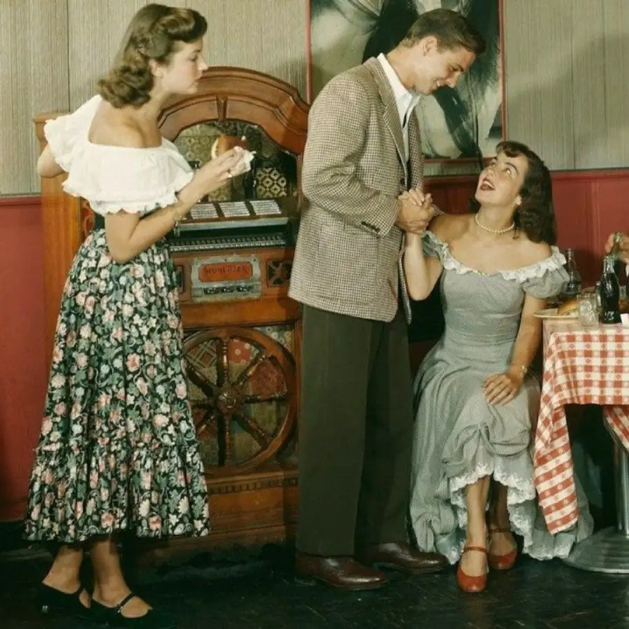 Three people in vintage 1950s attire are near a jukebox. A standing man holds hands with a seated, smiling woman, while another woman in a floral skirt stands nearby, watching. A checkered tablecloth is visible.