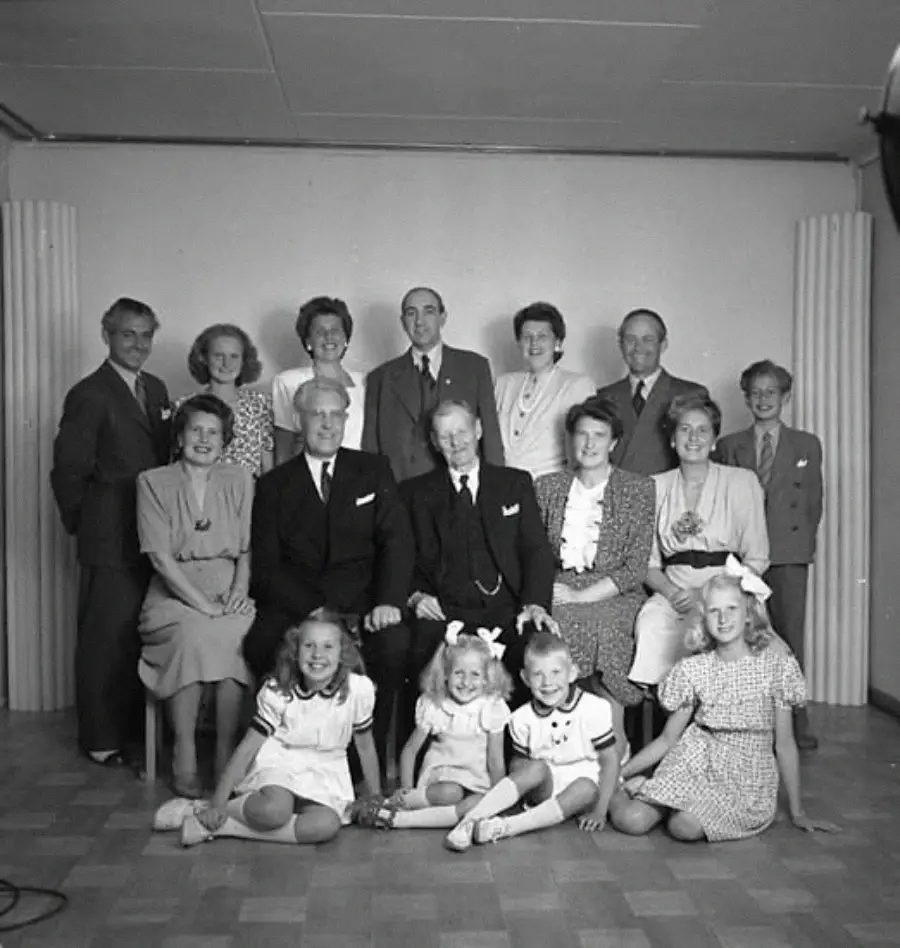 Black-and-white photo of a formal family group: eight adults standing, four adults seated, and four young girls sitting cross-legged on the floor, all smiling and dressed in 1940s-style clothing.