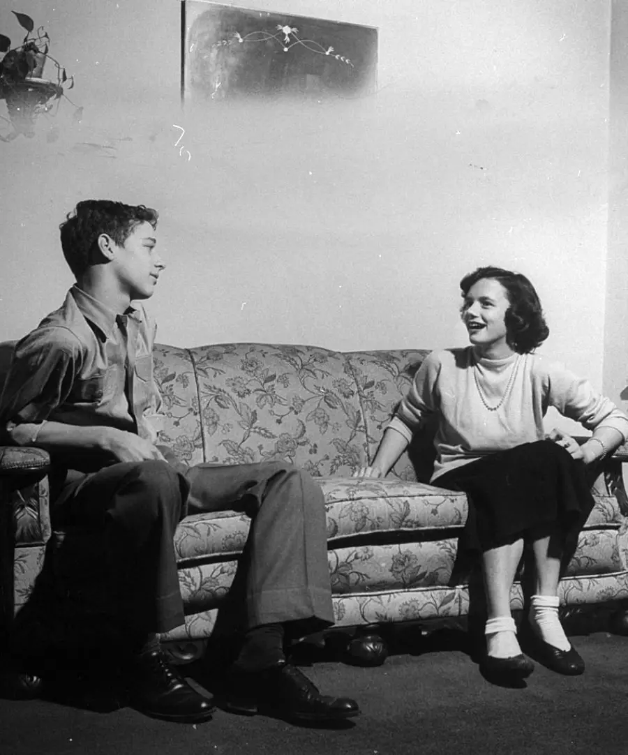 A young man and woman sit on a floral sofa in a living room, facing each other and smiling. A painting hangs on the wall behind them.