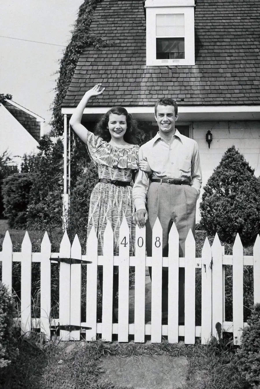 A smiling woman waves while standing next to a man in front of a white picket fence and a house with the number 408. They appear happy, holding hands, and are dressed in vintage clothing. Shrubs and a sloped roof are visible behind them.