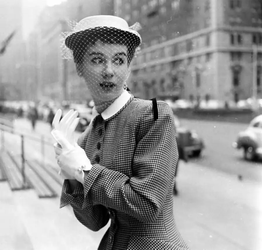 A woman in vintage attire, including a hat with a veil and gloves, poses on a city street. Cars and tall buildings are visible in the blurred background.