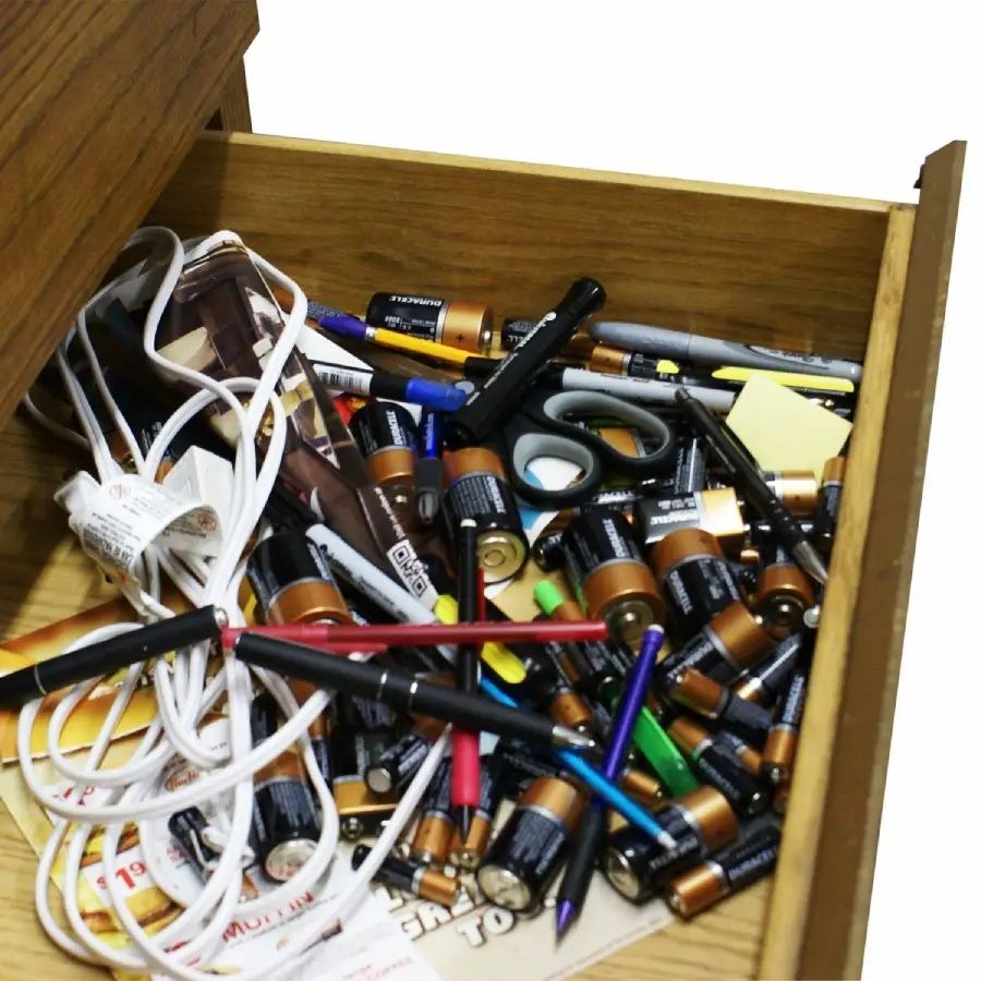 An open wooden drawer filled with loose batteries, pens, scissors, cables, sticky notes, a tape measure, and various small office items in a cluttered arrangement.