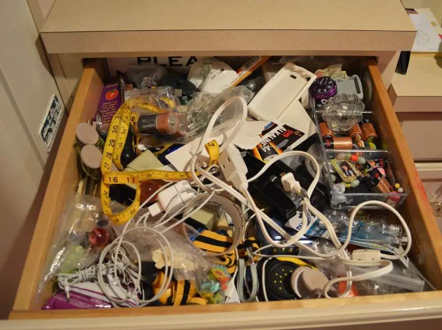 A cluttered drawer filled with tangled cords, batteries, tape measures, pens, plastic bags, tools, and assorted household items.