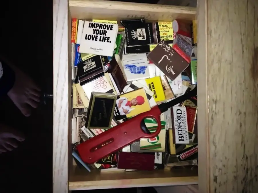 A wooden drawer filled with various colorful matchbooks, a red lighter, and a magnifying glass. Two feet are partially visible at the bottom left corner of the image.