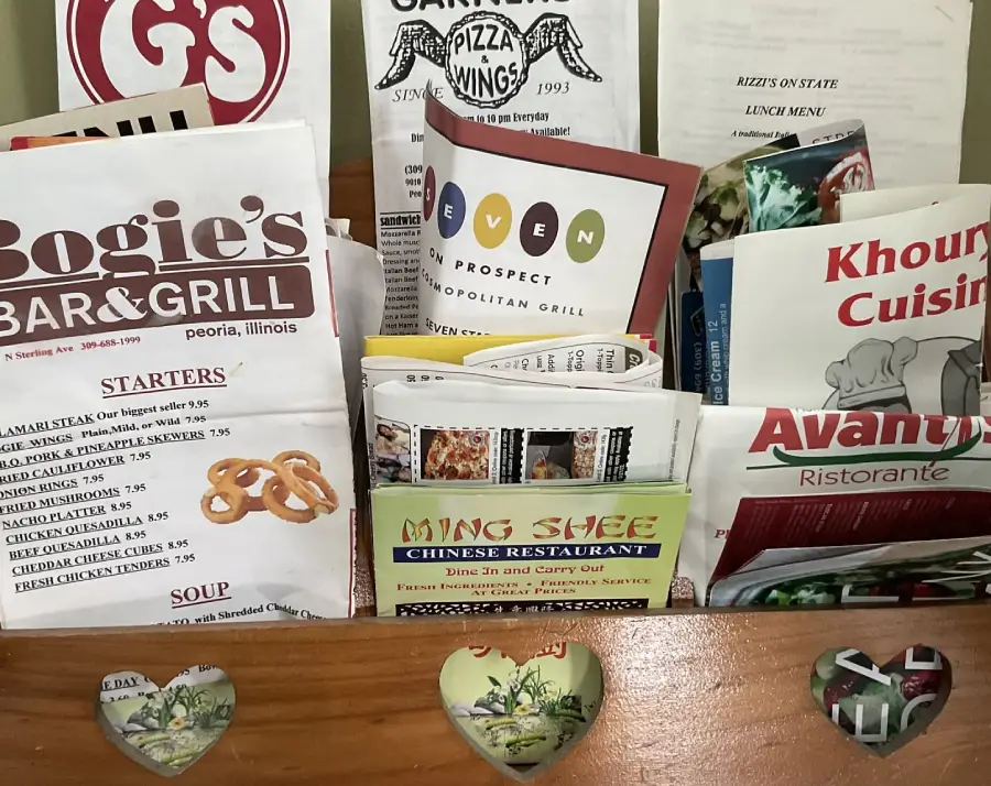 A wooden holder with heart cutouts filled with various restaurant menus, including Bogie's Bar & Grill, Ming Shee Chinese Restaurant, Khouri's Cuisine, and others.