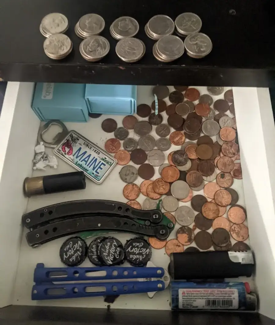 An open drawer containing stacks of coins, scattered loose change, a Maine keychain, two utility knives, bottle caps, a lighter, and a few small boxes.