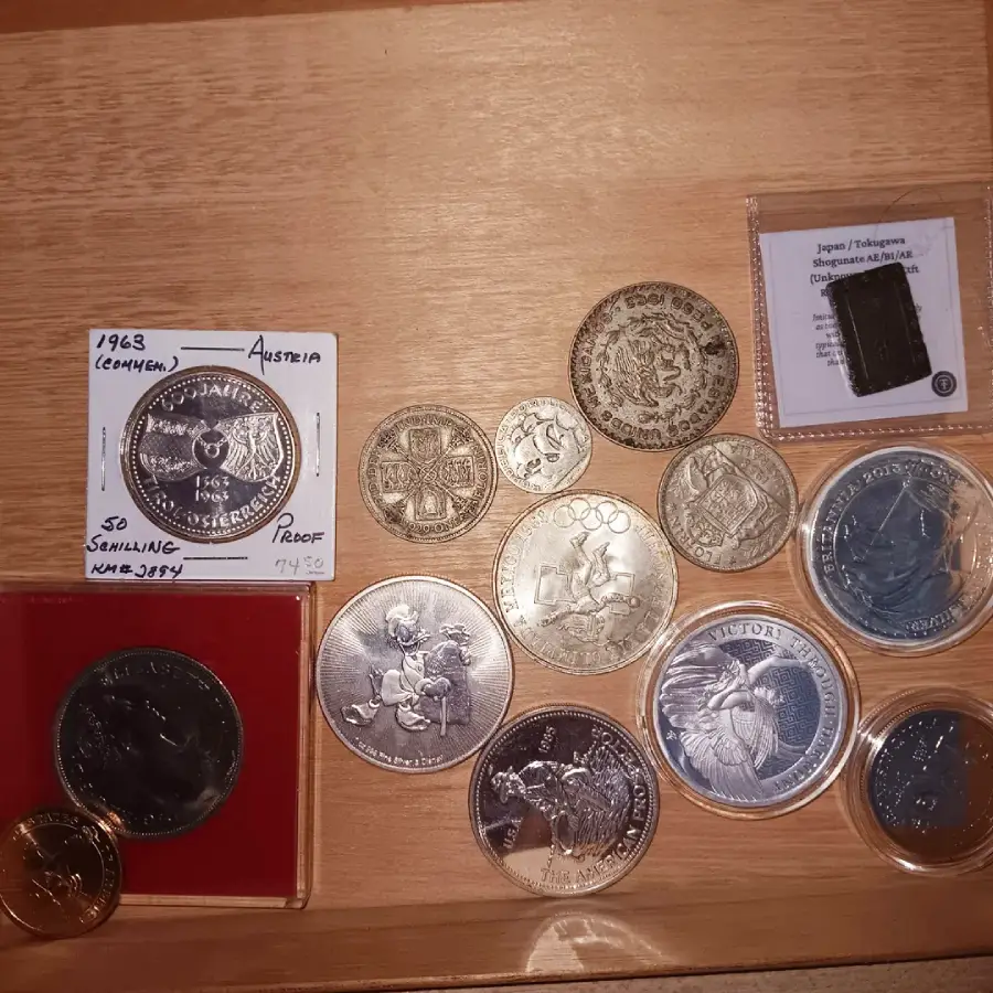 Various collectible coins, including commemorative and proof coins, are arranged on a wooden surface. Some coins are in holders or cases, with labels indicating origin, year, and details.