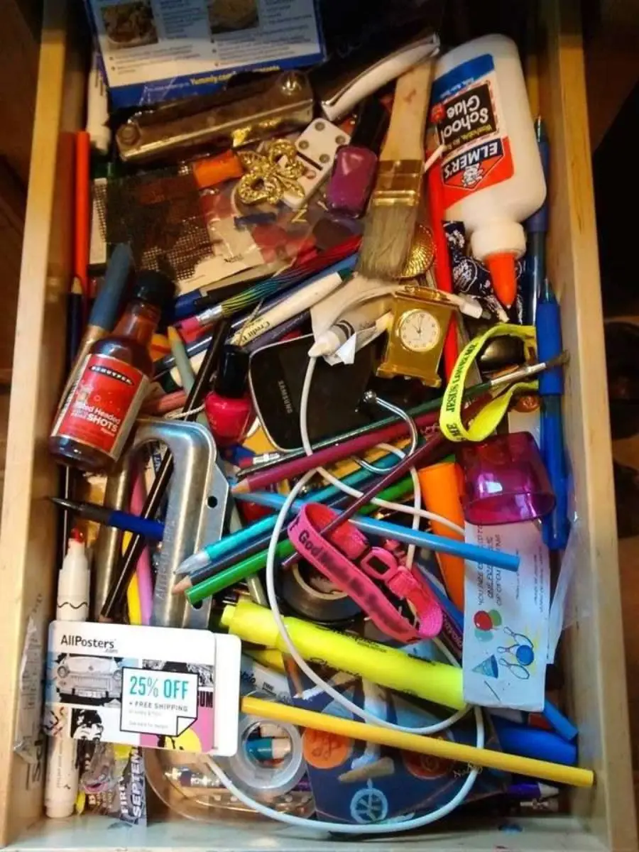 A messy drawer filled with various items including pens, glue, scissors, keys, a paintbrush, a watch, a lighter, coupons, and other miscellaneous objects piled together.