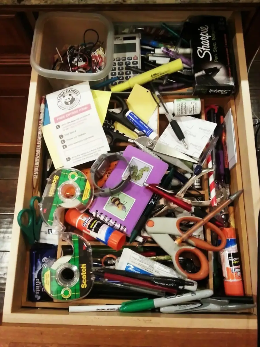 A cluttered drawer filled with various office supplies, including scissors, tape, glue sticks, pens, pencils, a calculator, sticky notes, markers, a container of small items, and miscellaneous papers.