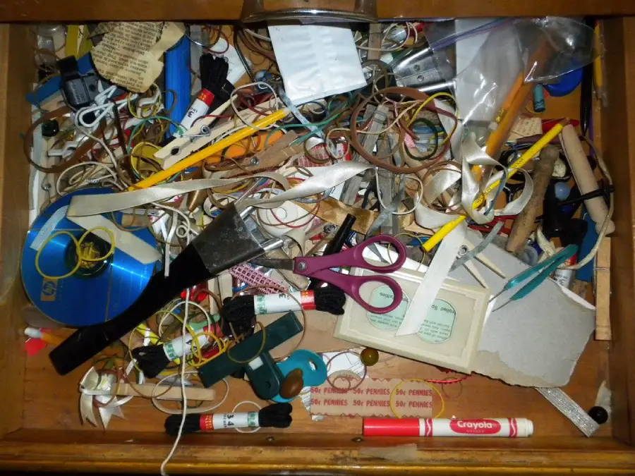 A cluttered drawer filled with assorted items including scissors, markers, spools of thread, rubber bands, ribbons, a paintbrush, and various small tools and objects.