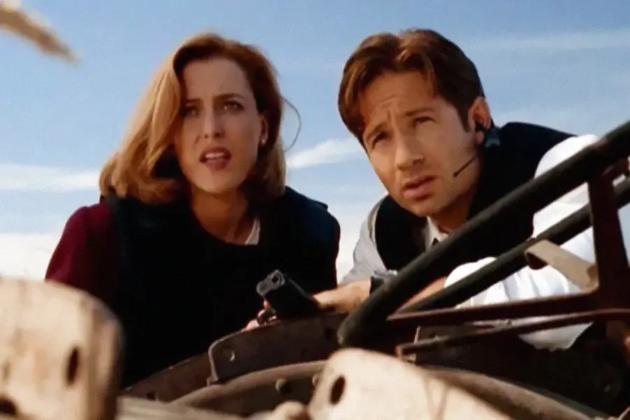 Two people wearing tactical vests crouch behind cover, looking intently ahead. One holds a gun and wears a headset. The sky is blue and clear in the background.