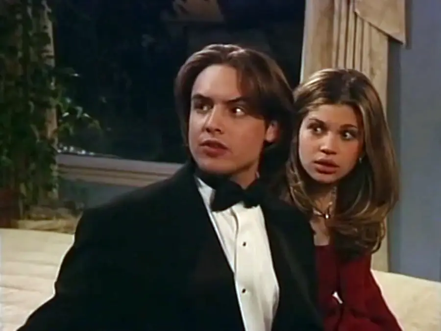 A young man in a black tuxedo sits beside a young woman with long brown hair, who appears surprised. They are indoors, with a window and curtains in the background.