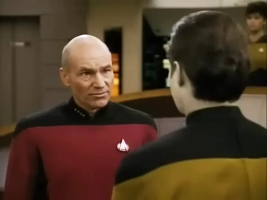 A man in a red and black uniform faces another person in a yellow and black uniform on a spaceship bridge, with a woman working at a console in the background.