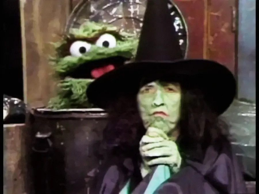 A person dressed as a witch with green skin and a black hat stands in front of a green puppet character popping out of a trash can.