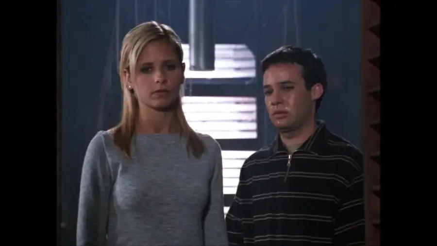 A blonde woman and a dark-haired man stand side by side in a dimly lit room, both looking serious and somber. Light filters in from a window behind them, casting shadows on the walls.