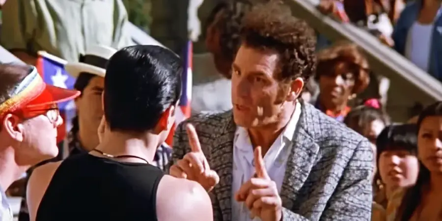 A man in a plaid jacket gestures with both index fingers raised while talking to another man in a black tank top, surrounded by a crowd outdoors.