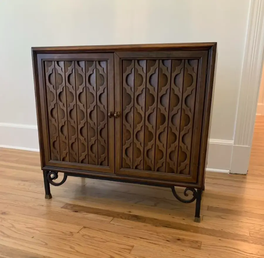 A small wooden cabinet with ornate carved doors and metal legs, placed on a light hardwood floor against a cream-colored wall.