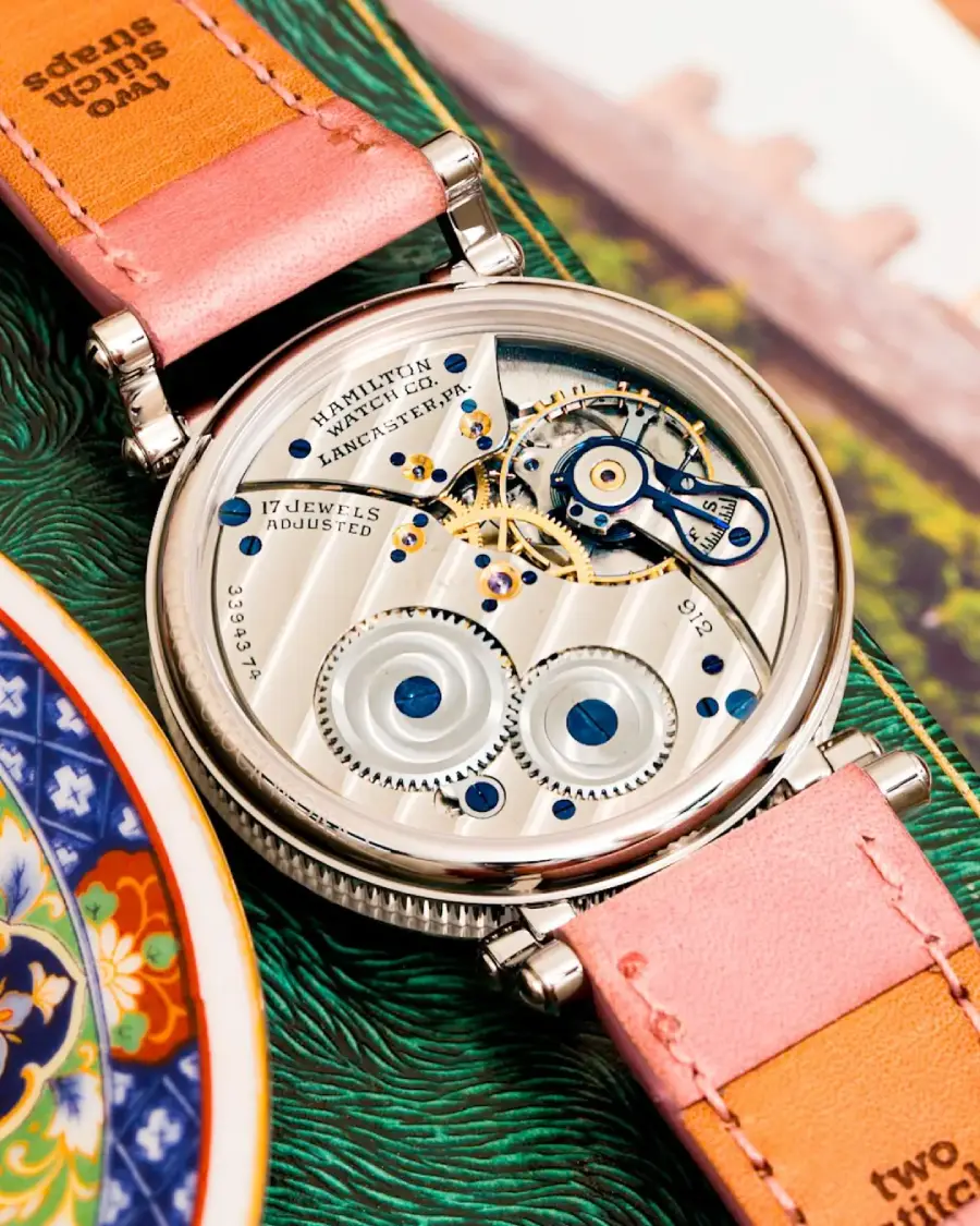 A close-up of a wristwatch’s intricate mechanical movement, displaying gears and components. The watch has a pink leather strap and is placed next to a colorful ceramic dish and a green textured surface.
