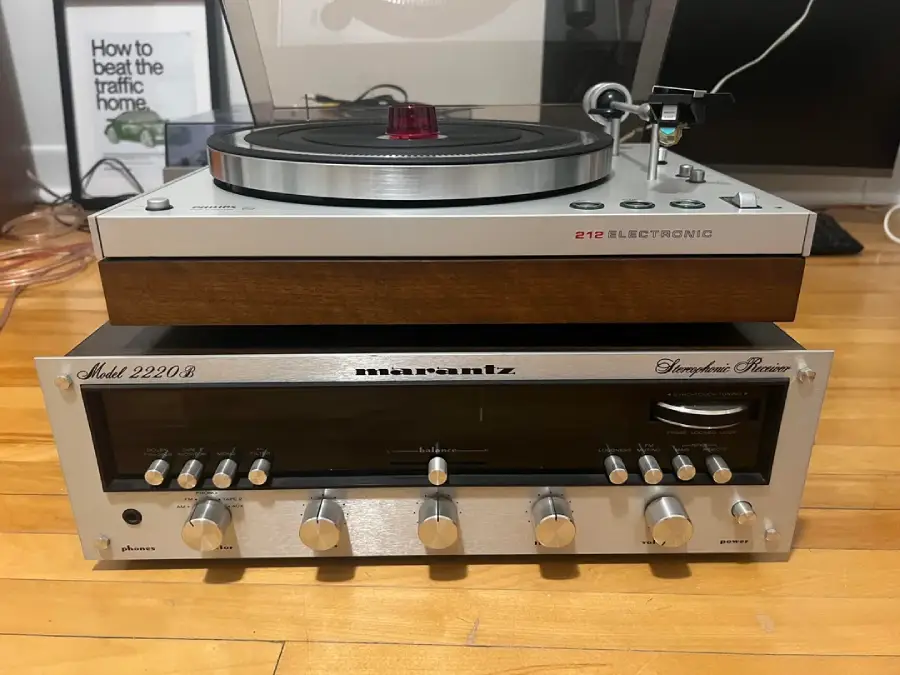 A turntable sits on top of a vintage Marantz Model 2220B stereo receiver, both placed on a wooden floor. A framed print and cables are visible in the background.