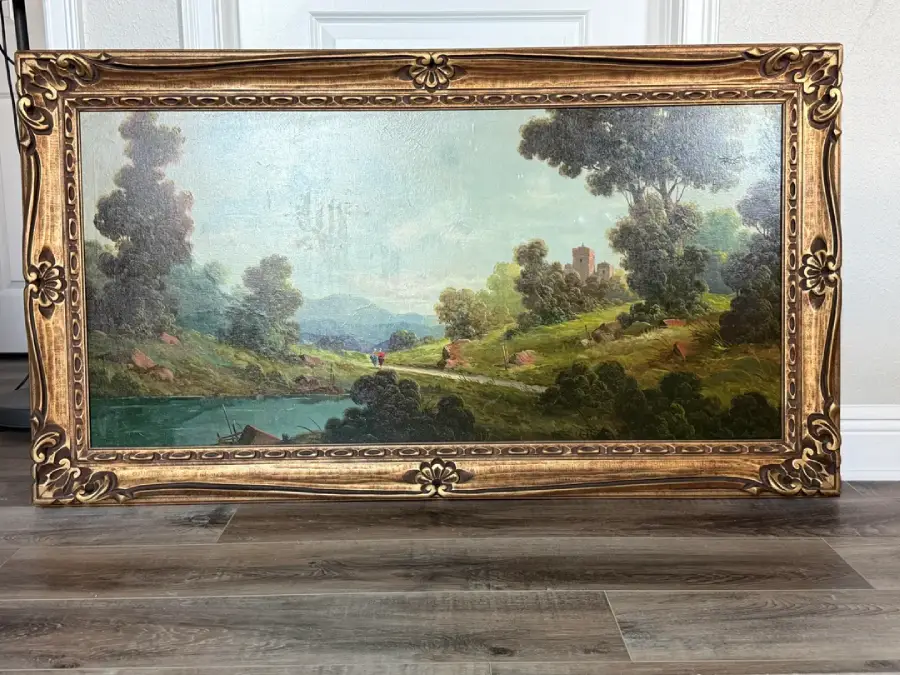 A framed landscape painting showing a river, trees, figures on grassy hills, and a distant castle, leaning against a white wall on a wood floor.