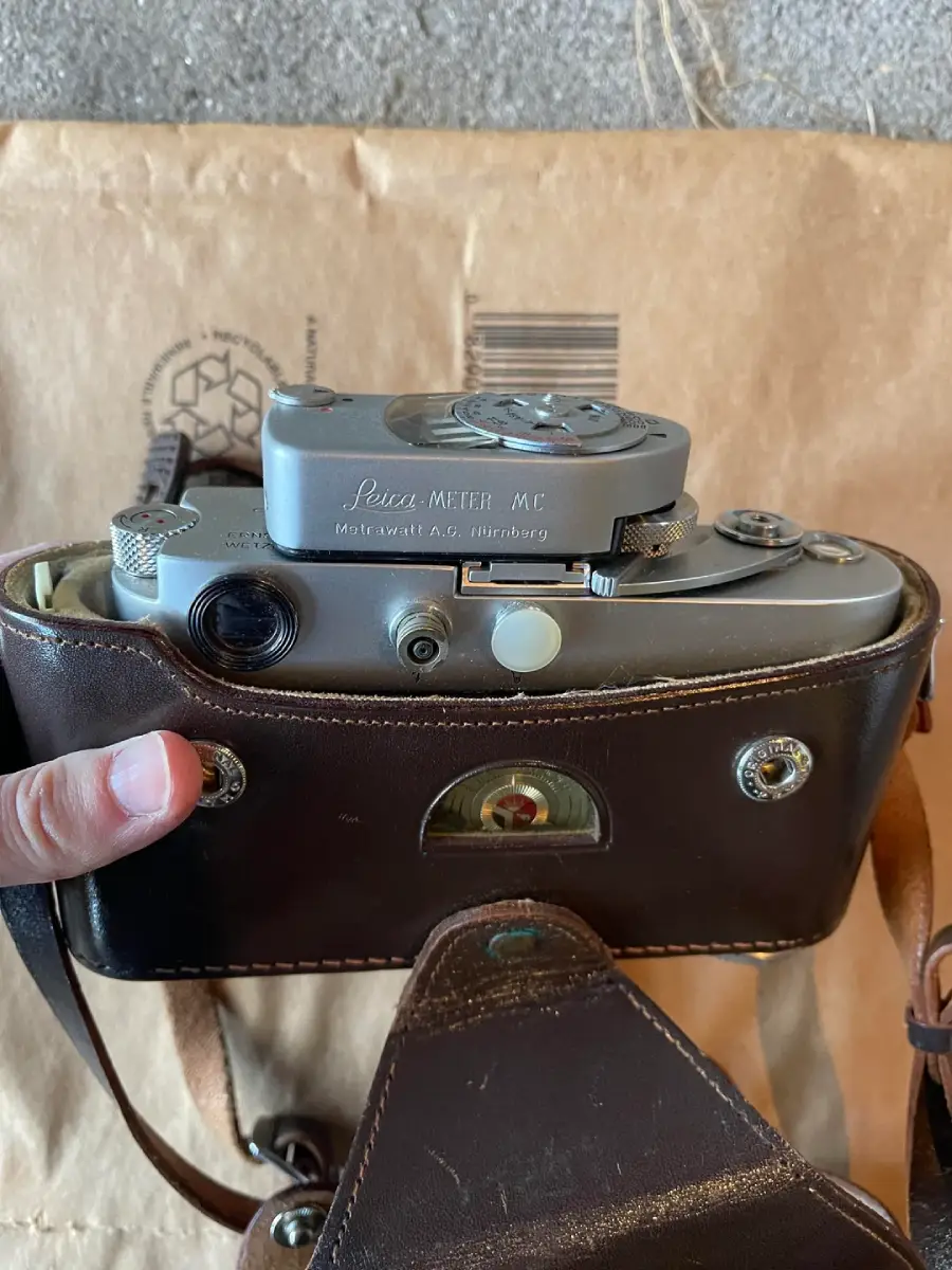 A hand holds a vintage Leica METER MC camera in a brown leather case, with part of a cardboard envelope visible in the background.