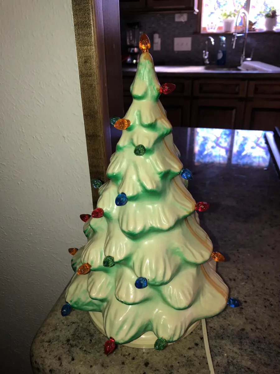 A ceramic Christmas tree with green accents and colorful bulbs sits on a kitchen counter, illuminated by natural light from a window in the background.