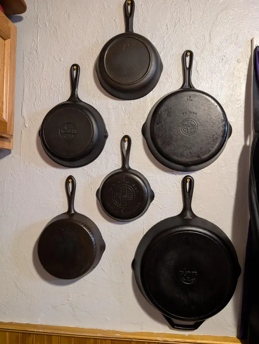 Six cast iron skillets of various sizes hang on a white wall in a neat arrangement, with a wooden cabinet partially visible on the left and wood paneling at the bottom.