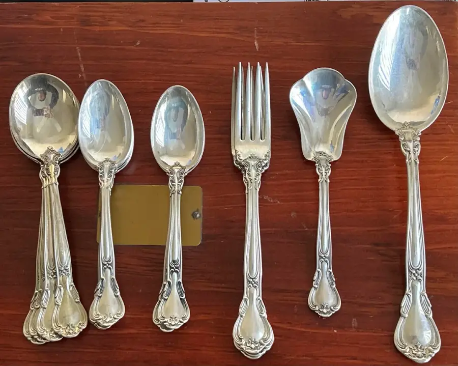 Six ornate silver spoons, one fork, and two serving utensils are arranged on a wooden surface with intricate designs on the handles.