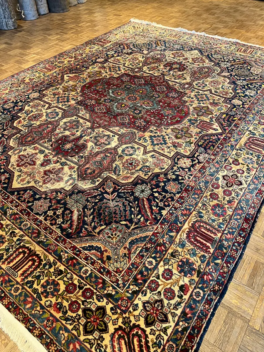 A large Persian rug with intricate floral and geometric patterns in red, navy, cream, and green colors, displayed on a wooden parquet floor.