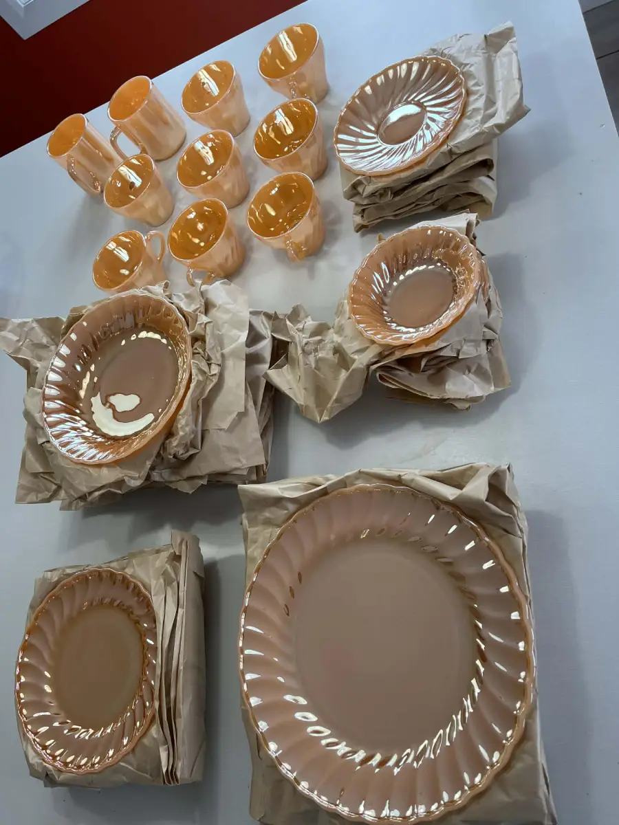 Peach-colored glass plates, bowls, and mugs are neatly arranged on a white surface, some stacked with brown paper between them.