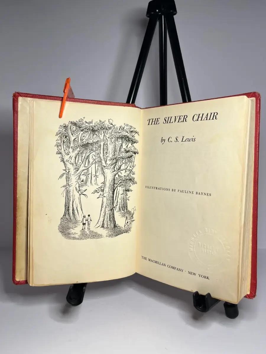 An open copy of “The Silver Chair” by C. S. Lewis on a black stand, showing the title page and an illustration of two small figures among tall, detailed trees. An orange bookmark sticks out from the top.