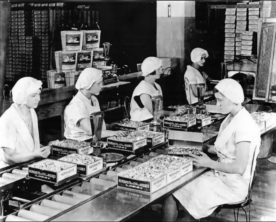 Black and white photo of women in uniforms and hairnets working on an assembly line, packing Hershey's chocolates into boxes inside a factory.