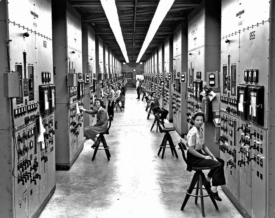 Black-and-white photo of women operating control panels in a long industrial room filled with gauges and switches, possibly from the mid-20th century. Several women sit on stools, monitoring equipment.