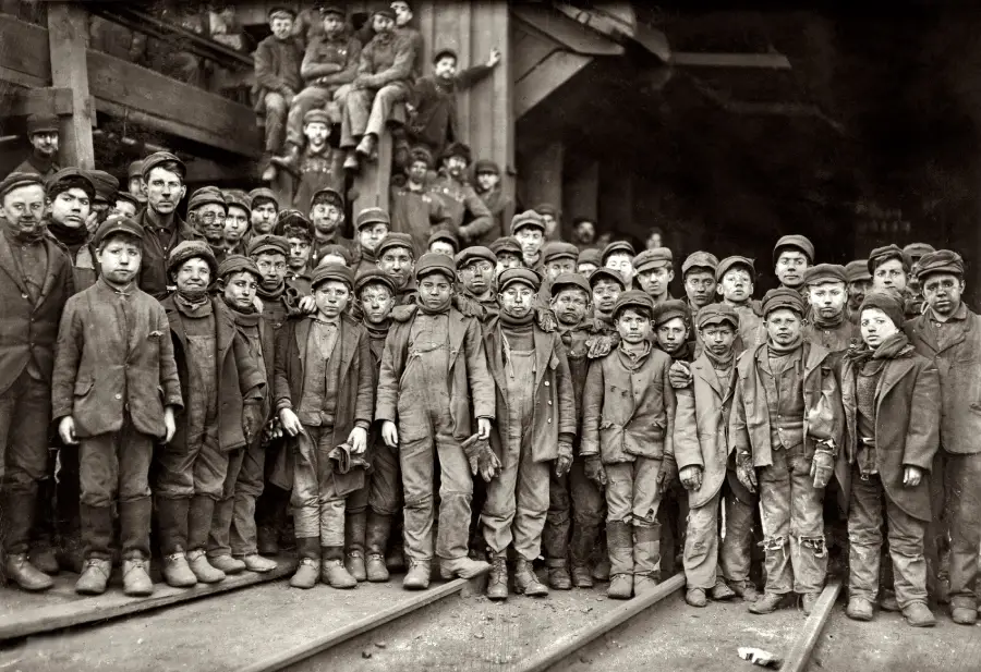 A large group of boys and young men in work clothing and caps stand close together on railroad tracks outside an industrial building, posing for a historical black-and-white photograph.