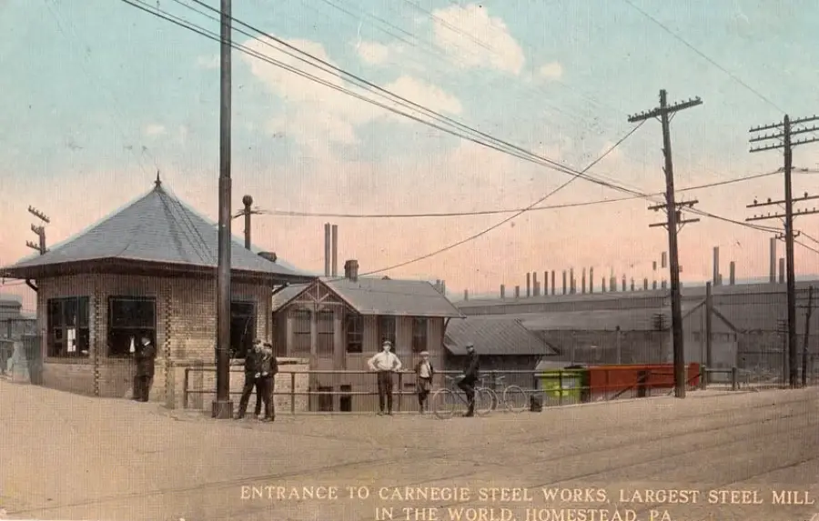 Historic photo of the entrance to Carnegie Steel Works in Homestead, PA, with workers standing near buildings and power lines; steel mill smokestacks visible in the background.