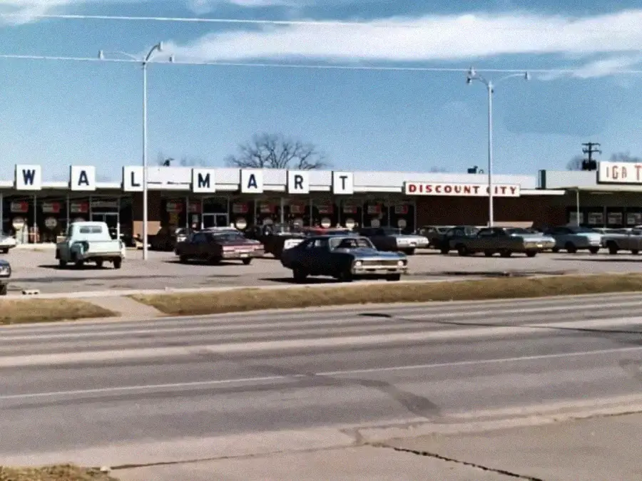A vintage photo shows an old Walmart Discount City store with classic cars parked in front, under a blue sky with scattered clouds. The store sign is partially visible.