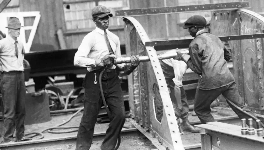 Two men work together using tools on a metal structure at an industrial site, while a third man stands nearby observing. All are wearing work clothes and hats, and the background shows industrial equipment and materials.