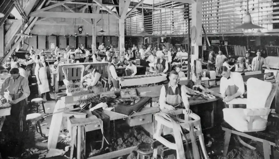 Black and white photo of a busy factory workshop with many workers assembling furniture at large tables; wood beams and tools are visible throughout the spacious, well-lit room.