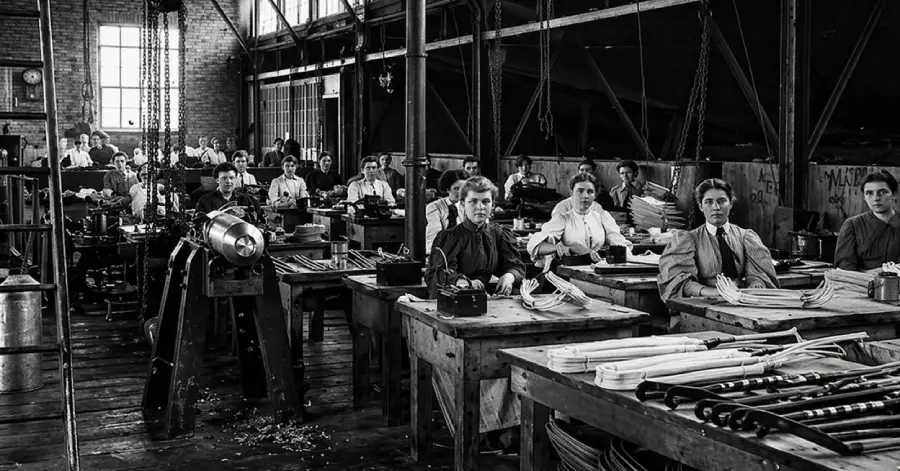 Black and white photo of women working at desks in a large factory or workshop, using machinery and assembling items, with natural light coming from tall windows.