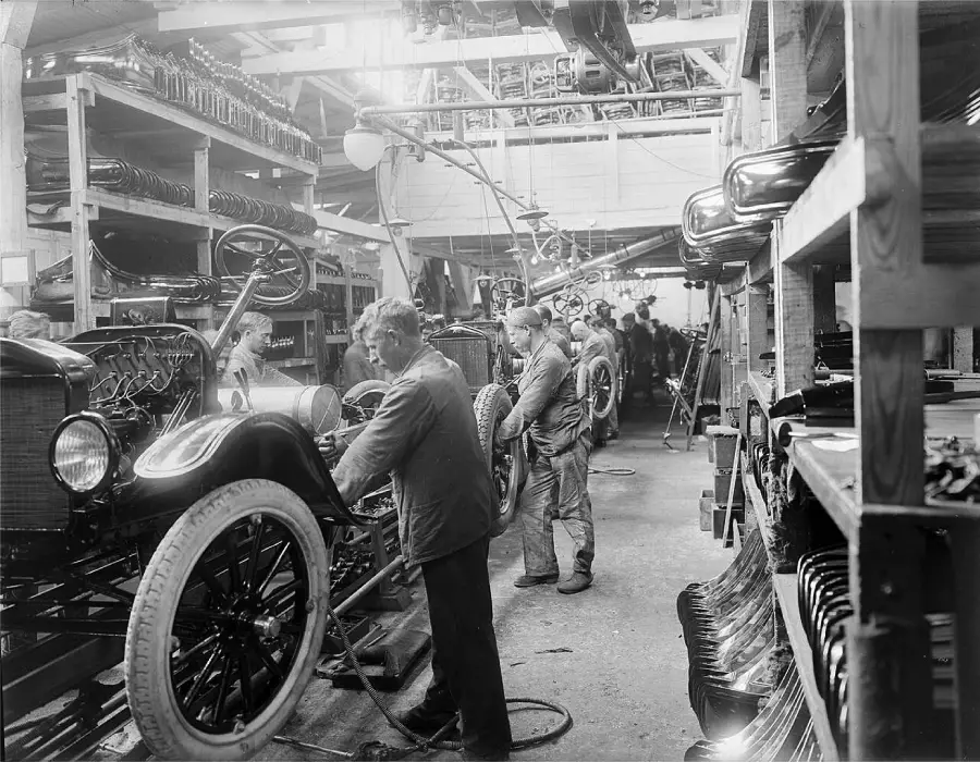 Workers assemble cars on an early 20th-century automotive assembly line inside a factory, with auto parts and tools visible on shelves and vehicles in progress.