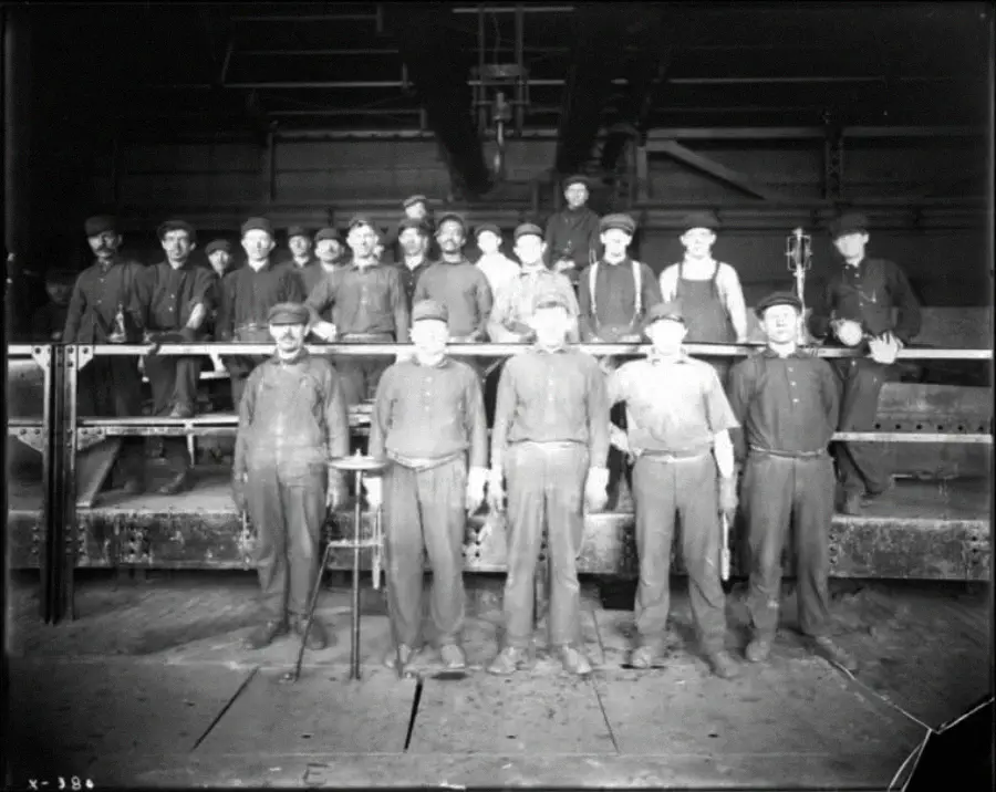 A black-and-white photo of about 20 factory workers standing in two rows on an industrial platform, wearing work clothes and hats, posing for the camera inside a large building.