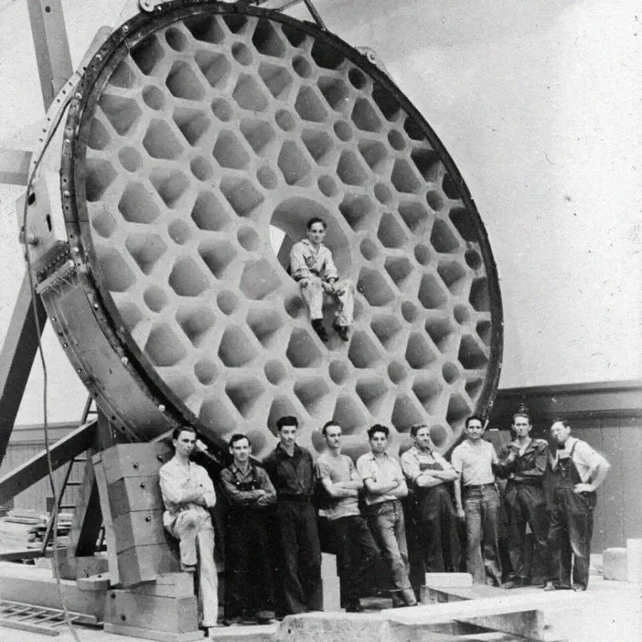 Eleven people stand in front of a giant honeycomb-patterned telescope mirror; one person sits in the central opening, highlighting the mirror's large size and intricate structure.