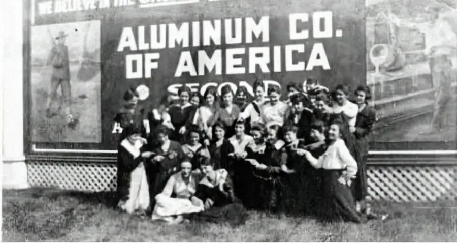 A group of women in early 20th-century clothing pose in front of a large "Aluminum Co. of America" sign outdoors. Most are smiling, and some are holding objects, likely at a company event or gathering.