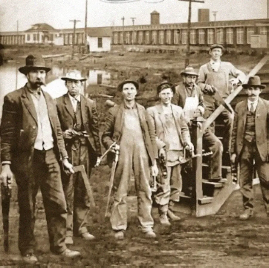 Historic black-and-white photo of seven men in work clothes and hats, some holding tools, standing outdoors near a wooden structure, with a large building and utility poles visible in the background.