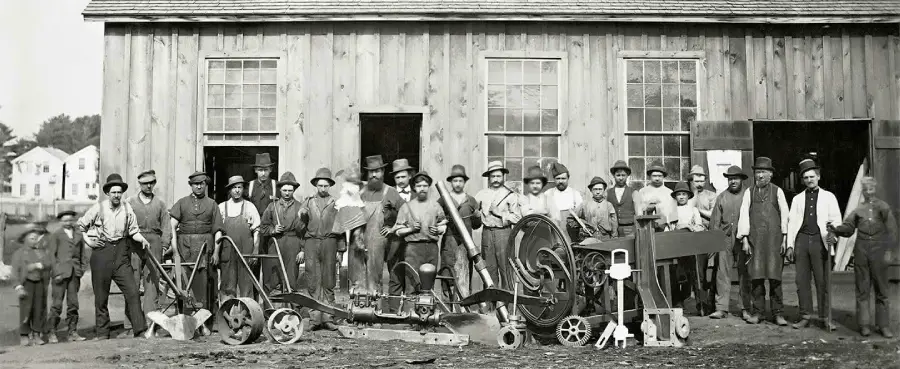 A group of men and boys stand in front of a wooden building with large windows, posing with various pieces of machinery and tools, likely from the late 19th or early 20th century.