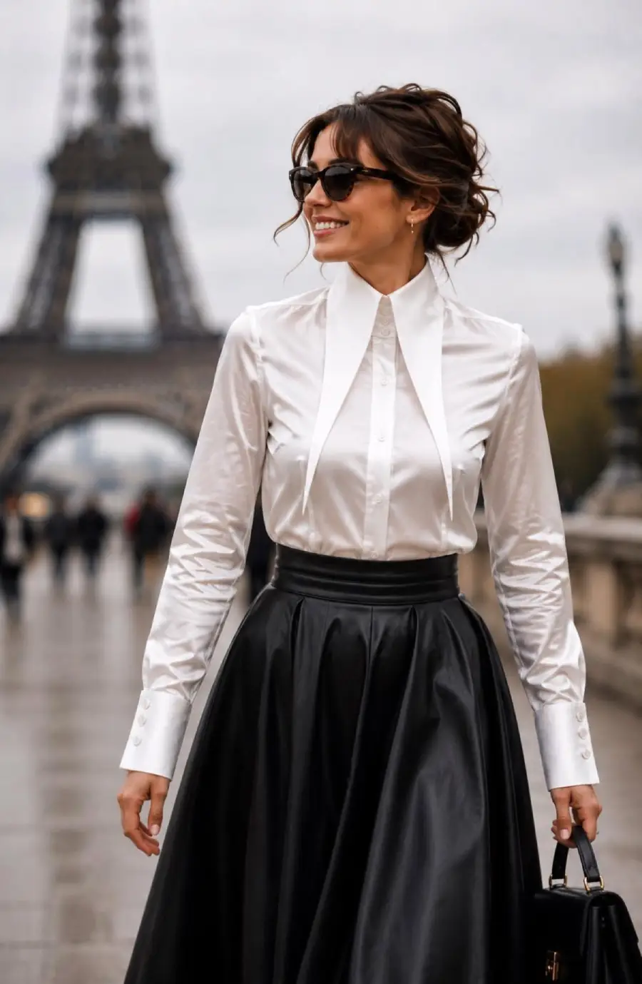 Woman in a white blouse and black skirt smiles while standing near the Eiffel Tower, holding a handbag. She wears sunglasses and her hair is styled up. The background is slightly blurred with people walking.